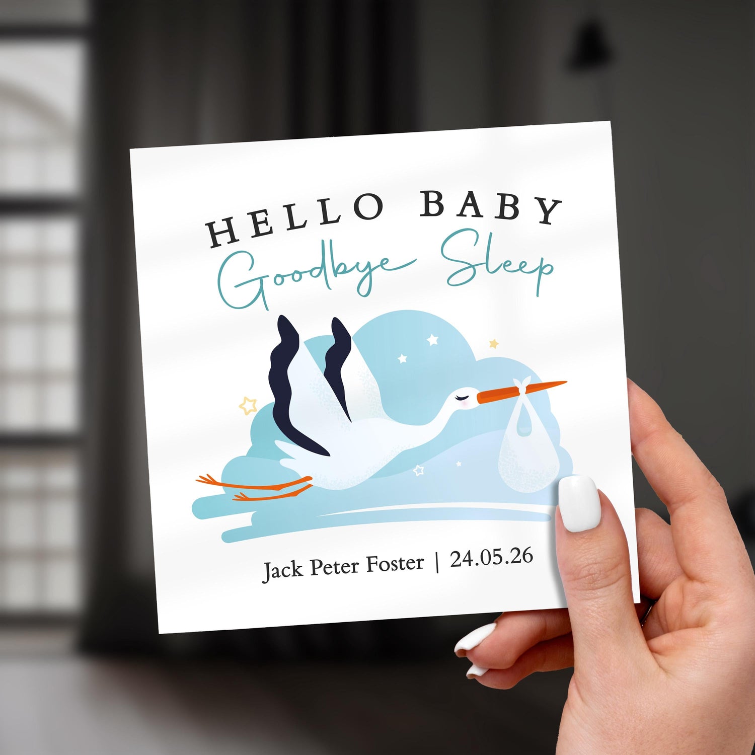 A modern new baby card featuring an illustrated stork carrying a baby bundle with the wording “Hello Baby, Goodbye Sleep”, photographed in hand against a softly blurred background.