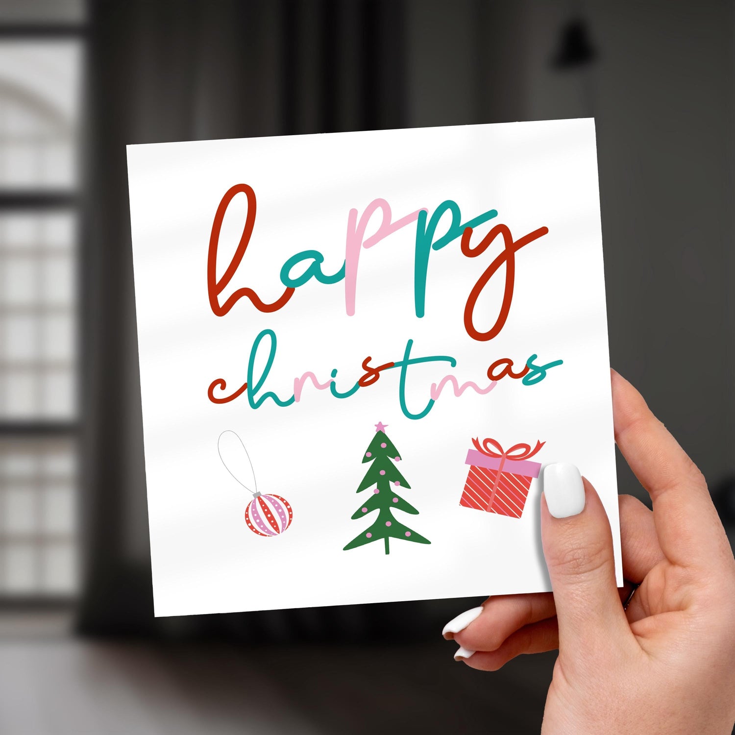 Festive Christmas card with colourful handwritten text ‘Happy Christmas’ and illustrations of a tree, present and bauble, held in hand indoors.