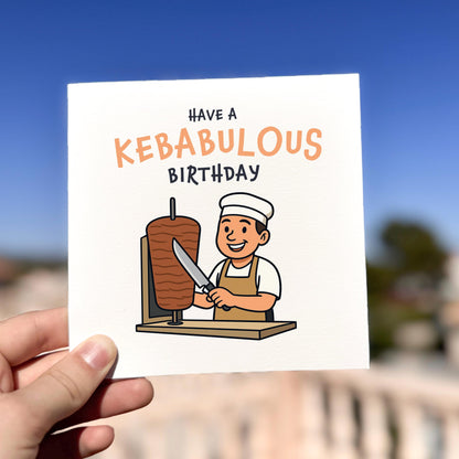 Card with cartoon chef and kebab, text 'Have a Kebabulous Birthday' held by a hand against a blurred outdoor background.