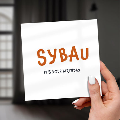 Hand holding a card with 'SYBAU It's Your Birthday' text against a blurred background