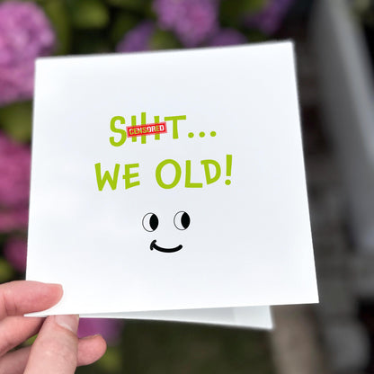 Hand holding a card with 'SHIT... WE OLD!' text and a smiley face, with a blurred floral background.
