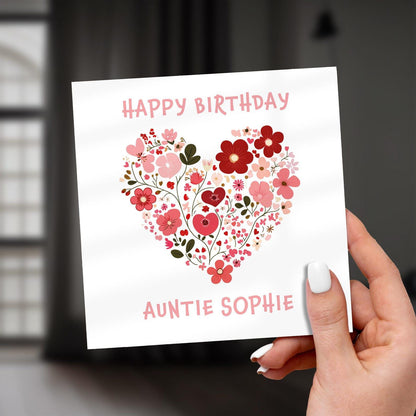 Hand holding a birthday card with floral heart design and text 'Happy Birthday Auntie Sophie' on a blurred background.