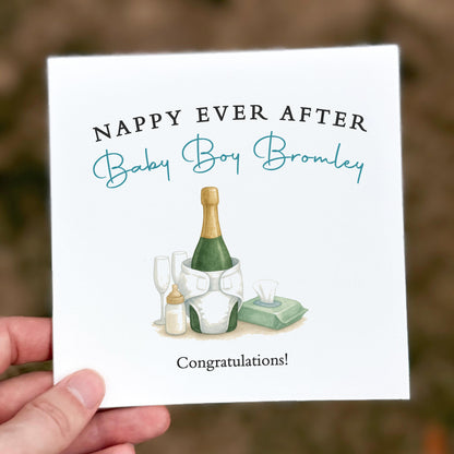 Greeting card with champagne bottle and baby items, text 'Nappy Ever After Baby Boy Bromley', held by a hand.