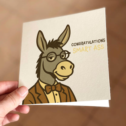 Greeting card with a donkey illustration and text 'Congratulations Smart Ass' held by a hand.