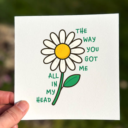 Card with a daisy illustration and text held by a hand against a blurred natural background