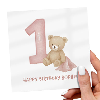 Hand holding a birthday card with a teddy bear and pink number '1' on a white background.