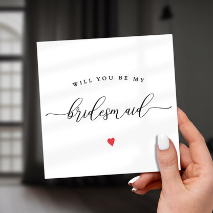 Hand holding a card with 'Will you be my bridesmaid?' text and a red heart on a white background.