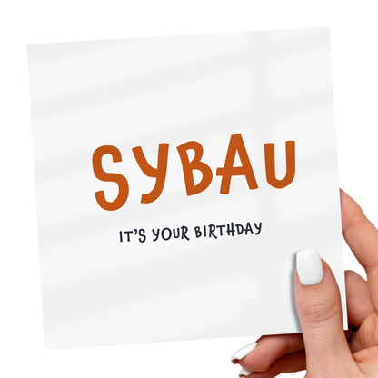 Hand holding a card with 'SYBAU IT'S YOUR BIRTHDAY' text on a white background