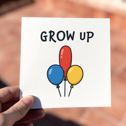 Card with colorful balloons and 'GROW UP' text held by a hand against a blurred background.