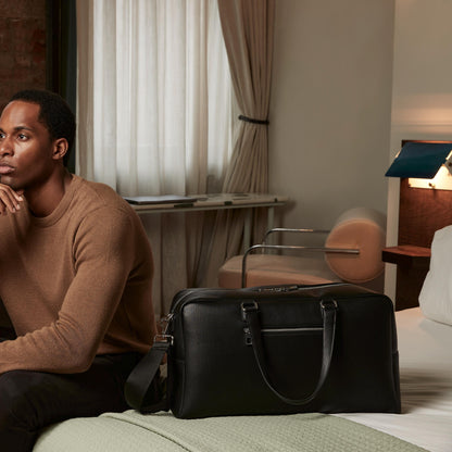 Man sitting on a bed with a black duffel bag in a room with curtains and a desk.