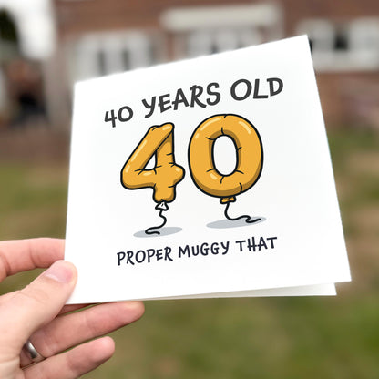 Hand holding a card with '40 Years Old 40 Proper Muggy That' text and cartoon balloons.