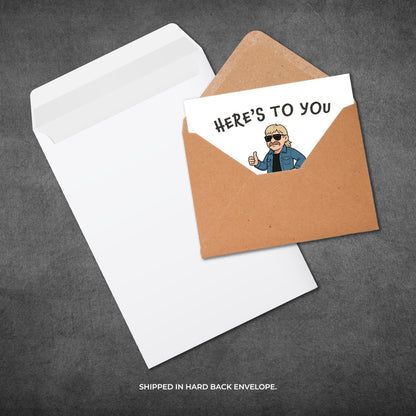 Greeting card with a cartoon character and 'Here's to You' text, shipped in a hard-back envelope on a gray background.