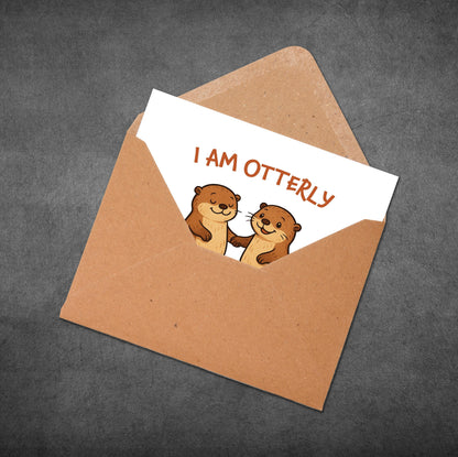 Card with two otters and 'I AM OTTERLY' text in an open brown envelope on a dark gray background