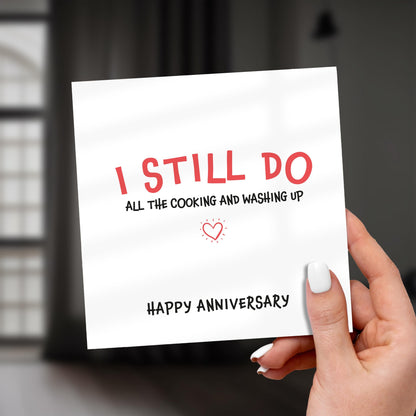 Card with 'I STILL DO all the cooking and washing up HAPPY ANNIVERSARY' text held by a hand.