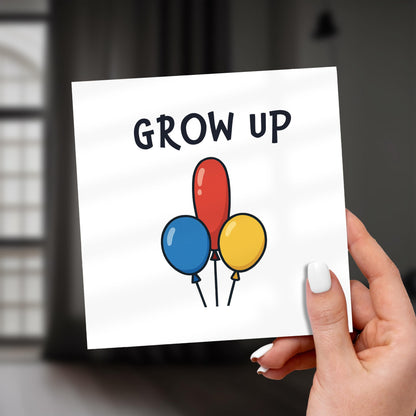 Hand holding a card with colorful balloons and 'GROW UP' text against a blurred background