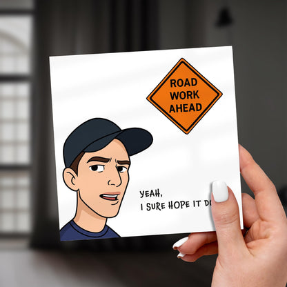 Hand holding a card with a cartoon character and 'Road Work Ahead' sign.