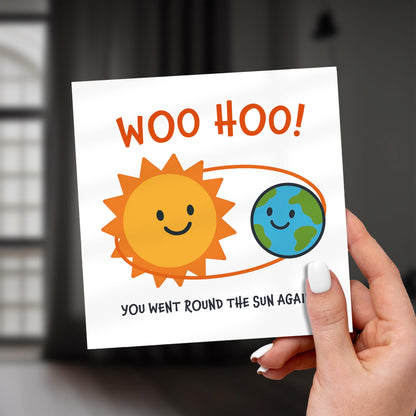 Hand holding a card with a sun and Earth illustration and text 'WOO HOO! You went round the sun again' against a blurred indoor background.