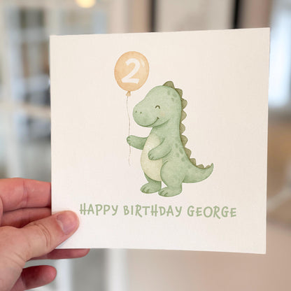 Birthday card with a dinosaur holding a balloon and text 'Happy Birthday George' held by a hand.