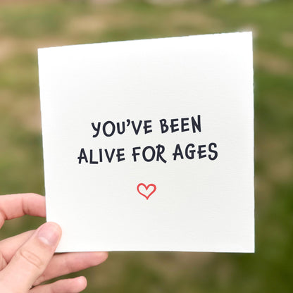 Hand holding a card with 'You've been alive for ages' text and a heart symbol against a blurred green background.