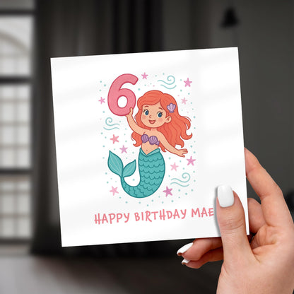 Hand holding a birthday card with a mermaid design and number 6 on a white background.