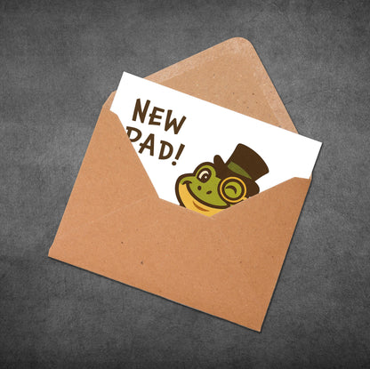 Greeting card with 'New Dad!' text and frog illustration in a brown envelope on a dark background
