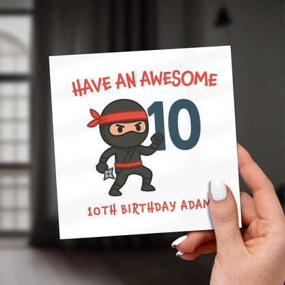 Hand holding a birthday card with a cartoon ninja and text 'Have an awesome 10 10th birthday Adam'.