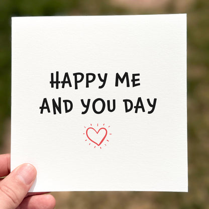 Hand holding a card with 'Happy Me And You Day' text and a heart symbol.