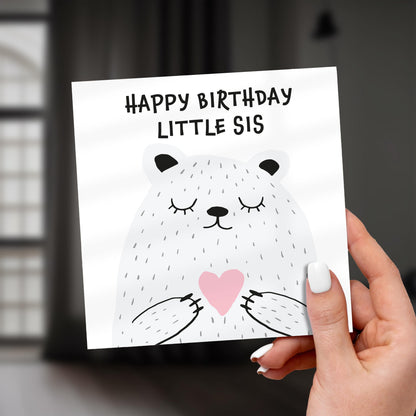 Hand holding a birthday card with a bear illustration and 'Happy Birthday Little Sis' text.