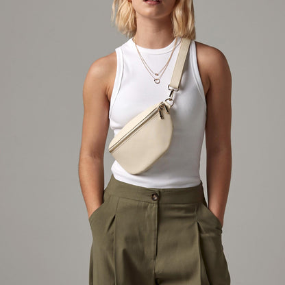 Person wearing a white sleeveless top and green pants with a beige crossbody bag on a gray background
