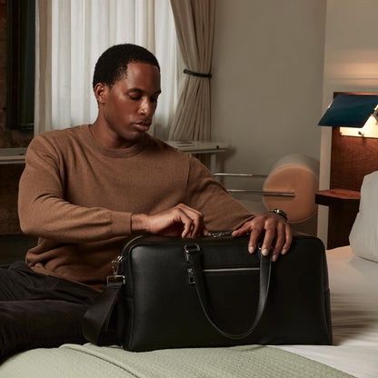 Man sitting on a bed holding a black leather bag in a bedroom setting.