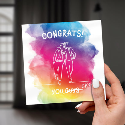 Hand holding a card with rainbow watercolor design and 'Congrats! You Guys Gay' text.