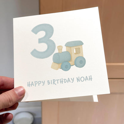 Hand holding a birthday card with a toy train and number 3 on it.