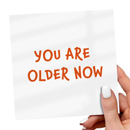 Hand holding a card with 'YOU ARE OLDER NOW' text on a white background
