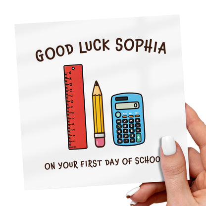 Card with 'Good Luck Sophia' on your first day of school text, ruler, pencil, and calculator illustrations held by a hand.