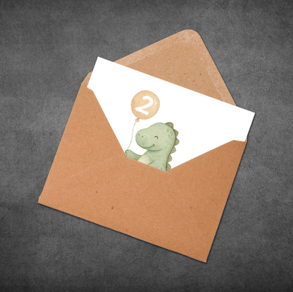 Brown envelope with a white card featuring a green dinosaur holding a number two balloon on a dark gray background.