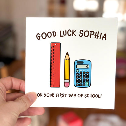 Card with 'Good Luck Sophia on your first day of school!' text and stationery items held by a hand.