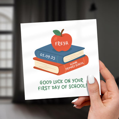 Hand holding a card with books and an apple illustration, featuring text 'Good luck on your first day of school!'.