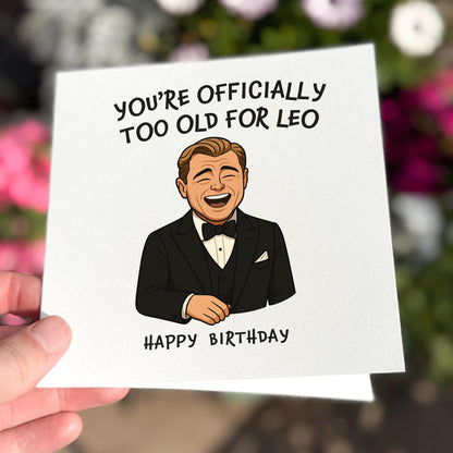 Birthday card with a cartoon of a man in a tuxedo and humorous text, held against a blurred floral background.