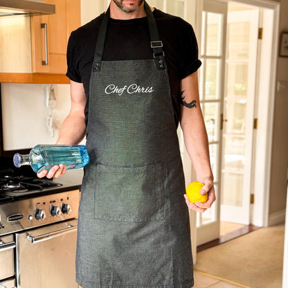 Person wearing a black apron with 'Chef Chris' text, holding a bottle and a lemon in a kitchen.