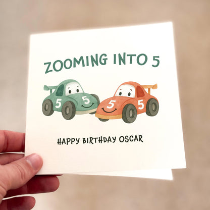 Birthday card with cartoon cars and text 'Zooming into 5, Happy Birthday Oscar' held by a hand.