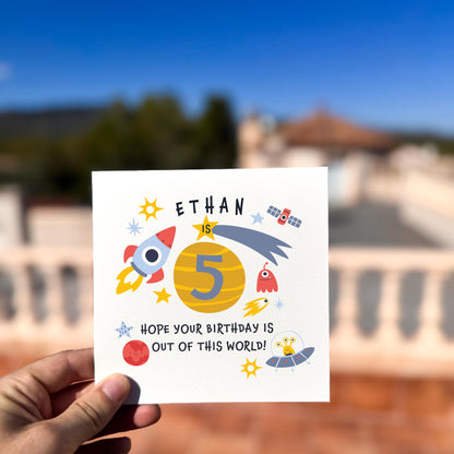 Hand holding a birthday card with space-themed design and text, outdoors.