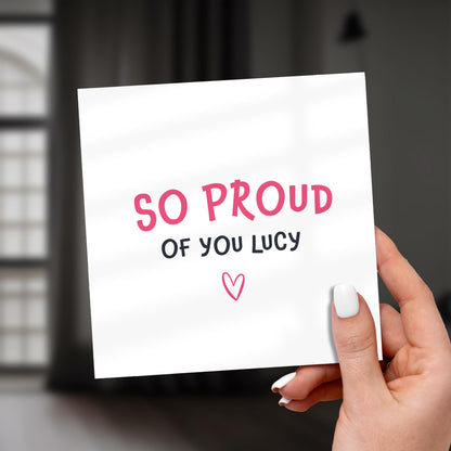 Hand holding a card with 'SO PROUD OF YOU LUCY' text and a heart symbol.