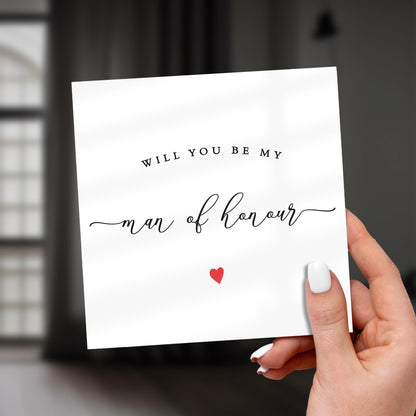 Hand holding a card with 'Will you be my man of honour' text and a red heart on a blurred background
