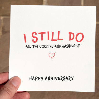 Card with 'I STILL DO all the cooking and washing up HAPPY ANNIVERSARY' text held by a hand.