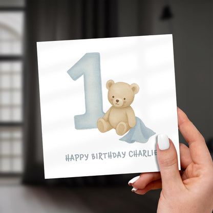 Hand holding a birthday card with a teddy bear and number '1' on a white background.