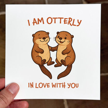 Card with cartoon otters and text 'I am otterly in love with you' held by a hand.