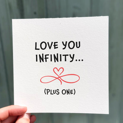 Card with 'Love you infinity... (plus one)' text and a red heart symbol held by a hand against a blurred background.