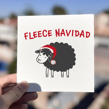 Card with a sheep wearing a Santa hat and 'Fleece Navidad' text held by a hand against a blurred outdoor background.