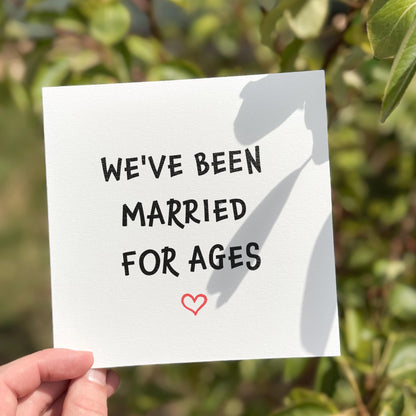 Hand holding a card with 'WE'VE BEEN MARRIED FOR AGES' text and a heart symbol against a green leafy background.