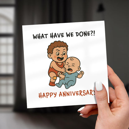 Card with cartoon illustration of two children and text 'What have we done?! Happy Anniversary!' held by a hand.
